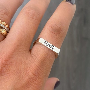 May include: A silver ring with the name "OLIVER" engraved in black block letters. The ring is rectangular with beveled edges and is worn on a finger. Another gold ring with small diamonds is also visible.