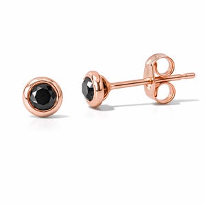 May include: A pair of rose gold stud earrings featuring black gemstones. The earrings have a bezel setting, with a round black stone in the centre of each earring. The earrings are displayed on a white background.