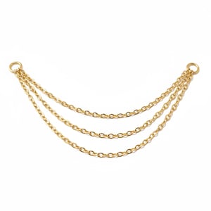 Puede incluir: Accesorio de cadena dorado con tres hebras paralelas. Cada hebra está compuesta por pequeños eslabones ovalados. El accesorio tiene un anillo en cada extremo para la fijación. Las cadenas cuelgan sobre un fondo blanco.
