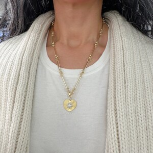 May include: A gold chain necklace with a heart-shaped pendant featuring a starburst design. The pendant is made of gold and has a textured surface.