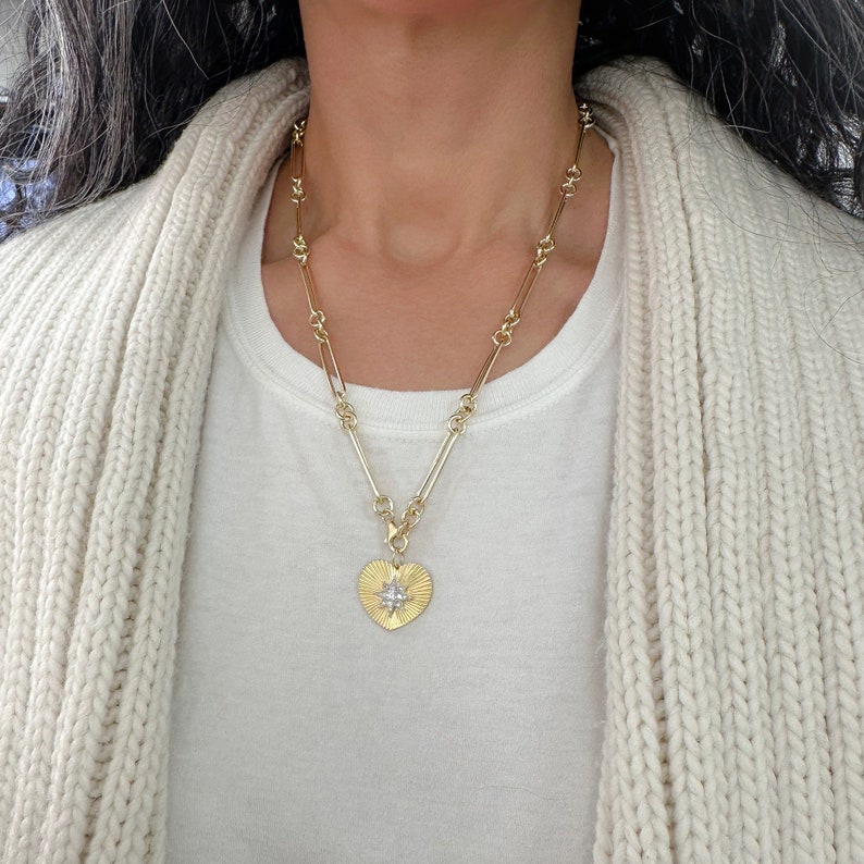 May include: A gold chain necklace with a heart-shaped pendant featuring a starburst design. The pendant is made of gold and has a textured surface.