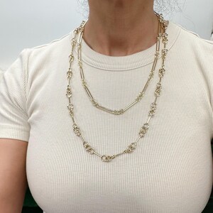May include: Two gold chain necklaces layered on a person's neck. The top necklace is a simple chain with rectangular links. The bottom necklace is a more intricate chain with round links and small bars connecting them.