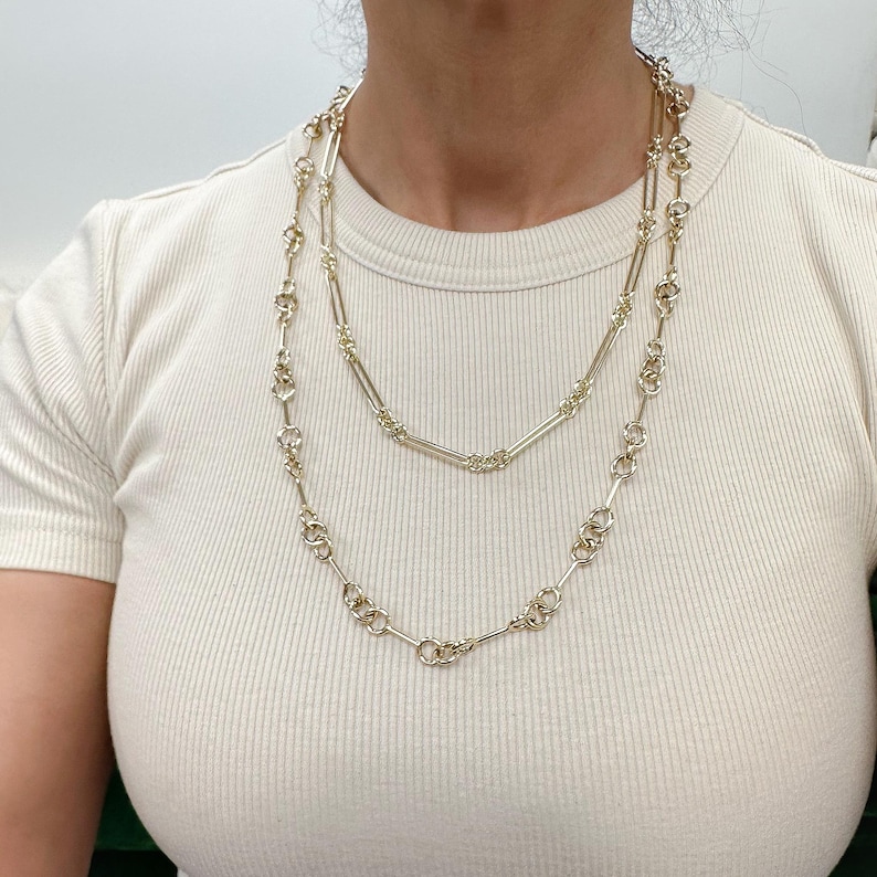 May include: Two gold chain necklaces layered on a person's neck. The top necklace is a simple chain with rectangular links. The bottom necklace is a more intricate chain with round links and small bars connecting them.