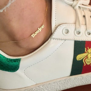 May include: A gold necklace with the name "Boujee" spelled out in cursive letters. The necklace is worn by a person wearing white sneakers with a green and red stripe and a bee design.