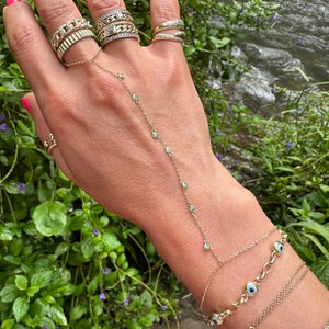 Könnte beinhalten: Eine goldene Handkette mit kleinen türkisfarbenen Steinakzenten, die mit einem Ring am Mittelfinger verbunden ist. Die Hand ist mit mehreren goldenen Ringen geschmückt, darunter Gliederketten-, Pavé- und glatte Ringe sowie ein Armband mit Nazar-Amuletten.