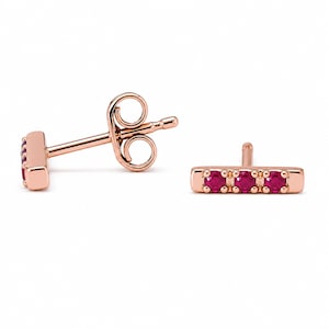 May include: A pair of rose gold bar stud earrings, each with three bright red gemstones. The earrings have a post and butterfly clasp. The design is simple and elegant, suitable for everyday wear or special occasions.