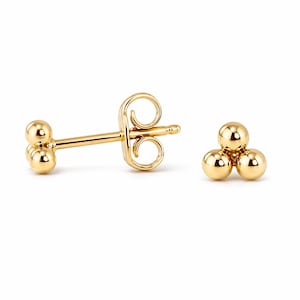 May include: A pair of gold-colored stud earrings. One earring features three small, round spheres arranged in a triangular cluster. The other earring has a post with two spheres and a butterfly backing. The earrings are set against a white background.