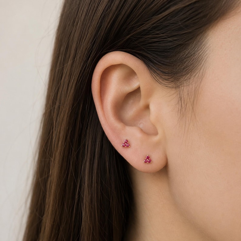 May include: Two small stud earrings, each featuring three tiny, vibrant pink stones arranged in a triangular cluster. The earrings are set in a metallic base and are positioned on the lower part of the earlobe, providing a subtle and delicate look.