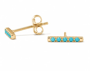 May include: A pair of gold-colored bar stud earrings. Each earring features a row of five small, bright turquoise stones. The earrings are displayed on a white background, showcasing their design and color.