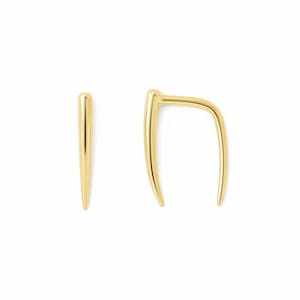 May include: A pair of gold-toned earrings consisting of one straight, tapered spike stud and one curved, U-shaped ear climber. Both pieces feature a smooth, polished metallic finish with a reflective surface and a minimalist, elongated design.