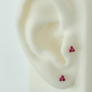 May include: Two stud earrings, each featuring three small, round, vibrant pinkish-red gemstones arranged in a triangular cluster. The stones are set in a metallic base, with one earring positioned on the tragus and the other on the earlobe.