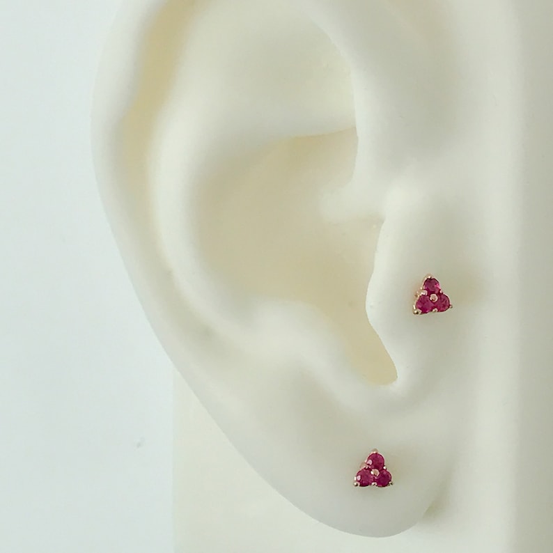 May include: Two stud earrings, each featuring three small, round, vibrant pinkish-red gemstones arranged in a triangular cluster. The stones are set in a metallic base, with one earring positioned on the tragus and the other on the earlobe.
