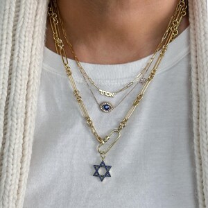 May include: A gold chain necklace with three pendants. The top pendant is a small gold charm with the word "TION" on it. The middle pendant is a blue evil eye charm. The bottom pendant is a gold Star of David charm with blue stones.