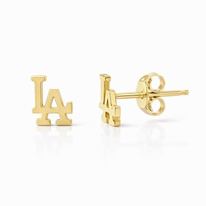 May include: A pair of gold-coloured stud earrings. Each earring is in the shape of the letters "LA", with a post and backing. The earrings are displayed on a white background.
