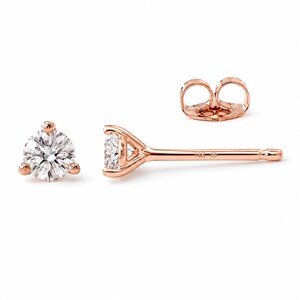 May include: A pair of rose gold stud earrings featuring round brilliant-cut clear stones held in three-prong settings. The posts are stamped with 14K and a small hallmark symbol. The earrings include matching rose gold butterfly-style backings.
