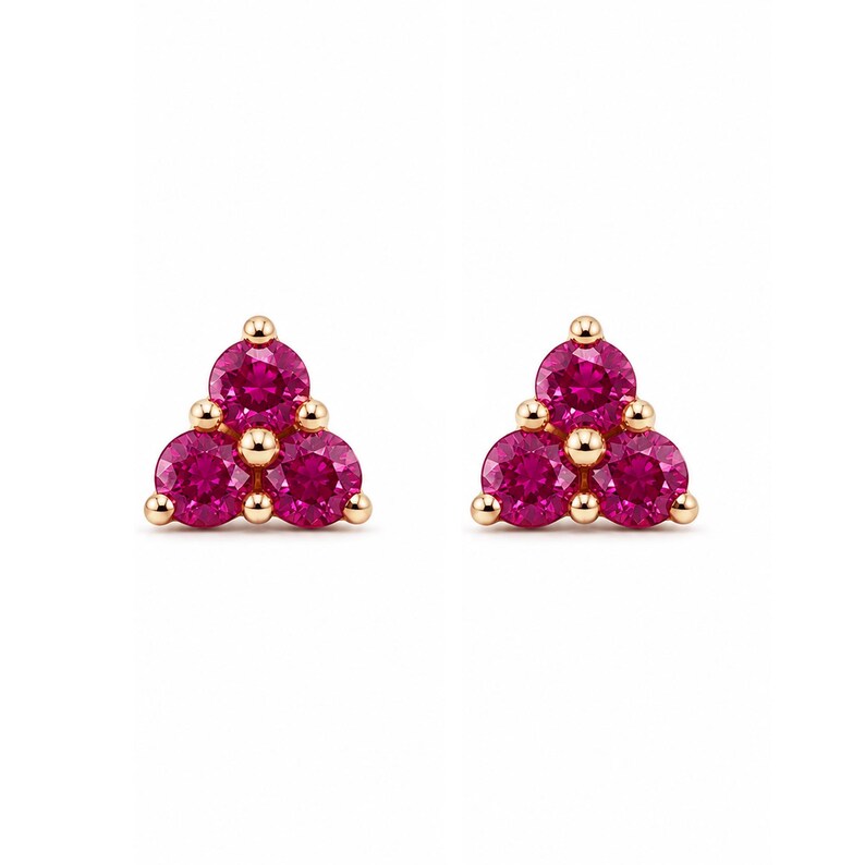 May include: A pair of stud earrings featuring three round, vibrant pink stones arranged in a triangular cluster. Each stone is set in a polished gold-tone metal setting with small, rounded prongs holding the gems securely in place.