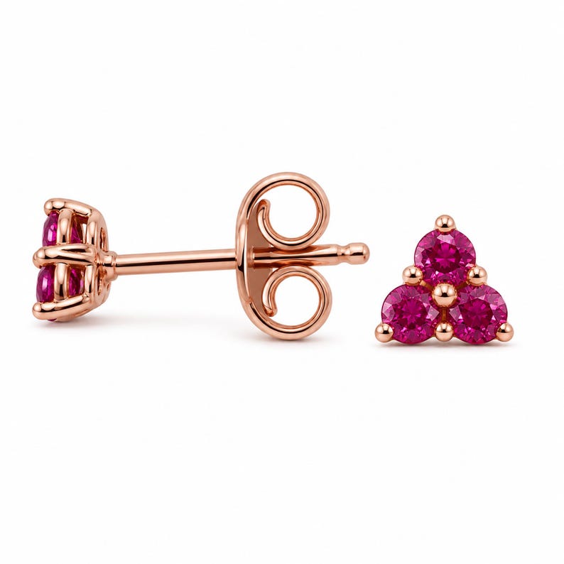 May include: A pair of rose gold stud earrings, each featuring three round-cut vibrant pink gemstones arranged in a triangular cluster. One earring is shown from the front, while the other is displayed from the side, revealing the post and back.
