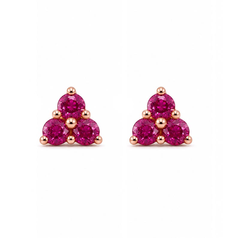 May include: A pair of stud earrings featuring three round-cut vibrant pink stones arranged in a triangular cluster. Each stone is set in a polished rose gold metal setting with small metallic prongs securing the gems in place.
