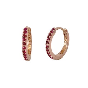 Może przedstawiać: Para kolczyków kółek z różowego złota. Każdy kolczyk jest inkrustowany rzędem małych, żywych czerwonych kamieni szlachetnych. Kolczyki są prezentowane na białym tle, podkreślając ich design.