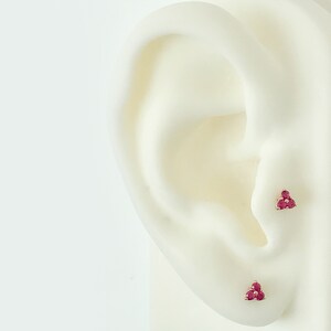 May include: Two stud earrings, each featuring three small, round, deep pinkish-red gemstones arranged in a triangular cluster. The earrings are set in a metallic base and are positioned on a white ear-shaped display model for demonstration.