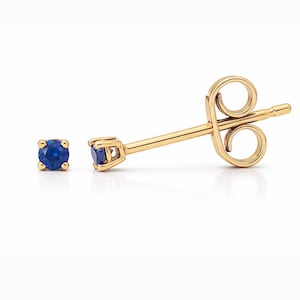 May include: A pair of gold stud earrings with a bright blue gemstone. One earring is in focus, showing the post and butterfly backing. The other earring is a single stud. The earrings are set against a white background.