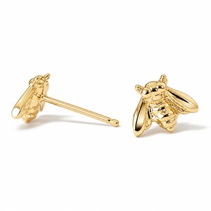 May include: A pair of gold-colored bee stud earrings. The earrings are detailed with wings, body segments, and antennae. The earrings are on a white background.