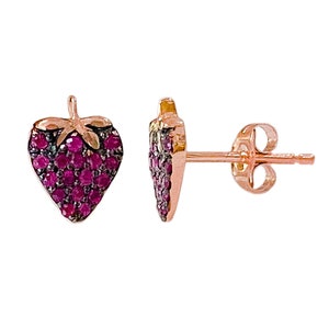 Puede incluir: Un par de pendientes de botón con forma de fresa. Los pendientes son de color oro rosa, con los cuerpos de fresa incrustados con pequeñas piedras redondas de color rubí. Las hojas de fresa son de un color más oscuro y contrastante.