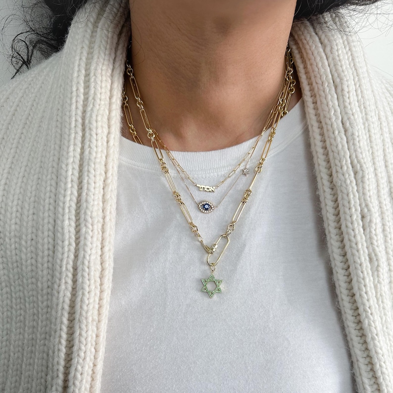 May include: A layered gold chain necklace with a small evil eye charm and a larger Star of David pendant. The Star of David pendant is green and has small clear stones around the edges.