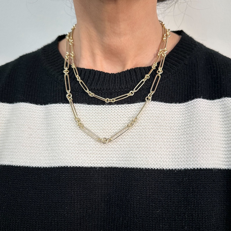 May include: Two gold chain necklaces layered on a person wearing a black and white striped jumper. The chains are made of linked oval shapes.