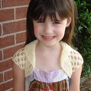 Op de afbeelding: Een jong meisje draagt een witte gehaakte bolero over een bloemenjurk. De bolero heeft een delicaat kantpatroon en is licht crème van kleur. Het meisje heeft lang bruin haar en glimlacht.