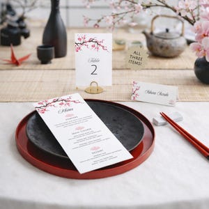 May include: A white menu card, a table number card labeled Table 2, and a place card labeled Sakura Suzuki, all featuring pink cherry blossom illustrations. The menu lists starters, main course, and dessert. A gold-toned stand holds the table card.