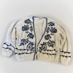 May include: Cream-coloured cardigan sweater with blue floral embroidery. The V-neck sweater features a button-up front and blue trim along the edges and sleeves. The floral design is repeated on the front and sleeves.