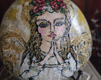 May include: A hand-painted river stone featuring an angel with long hair, a floral crown, and praying hands. The angel has gold glitter wings and a gold halo. The background is painted in shades of yellow and white, with black outlines defining the figure.