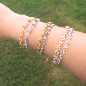 Puede incluir: Varios brazaletes de cuentas con un diseño de flor de margarita. Los brazaletes son de color dorado, plateado y blanco. Los brazaletes se llevan en un brazo, con un fondo de hierba verde.