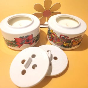 Può includere: Due portacandele in ceramica bianca con disegni festivi natalizi, tra cui un fiocco rosso e strumenti musicali. I portacandele hanno coperchi bianchi abbinati con un design a forma di bottone. Una decorazione floreale in legno è sullo sfondo.