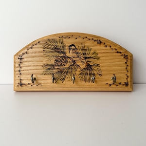 May include: A semi-circular wooden wall-mounted key holder featuring a wood-burned illustration of a small bird perched on a pine branch with a pinecone. The natural grain wood piece includes five metal hooks attached along the bottom edge.