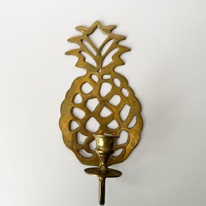May include: A brass pineapple-shaped wall sconce. The sconce features a detailed cutout design and a small cup to hold a candle. The pineapple's crown is at the top, and the candle holder is centered.