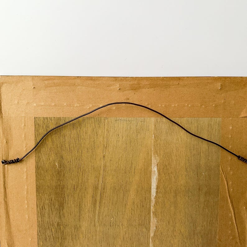 May include: Back of a framed artwork, showing the wooden frame, cardboard backing, and a black wire for hanging. The wood grain is visible on the frame and backing, with a neutral color palette.