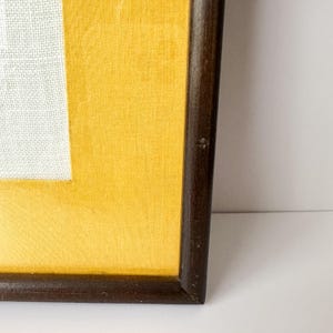 May include: A framed artwork featuring a white rectangular textile on a yellow background. The frame is made of dark brown wood. The artwork is displayed against a white background.