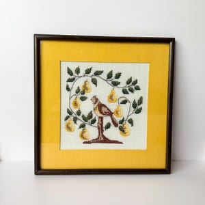 May include: Framed cross-stitch artwork featuring a bird perched on a stand amidst a pear tree. The embroidery showcases yellow pears, green leaves, and a brown bird against a cream background. The frame is dark brown with a yellow mat.
