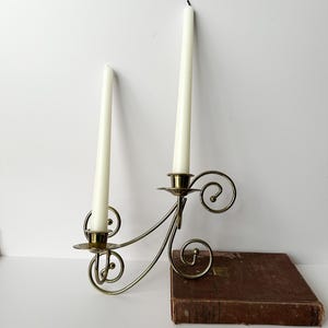 May include: A brass wall-mounted candle holder with two arms featuring curved scrollwork designs. Two off-white taper candles are inserted into the circular brass cups. The metal fixture has a polished finish and a symmetrical, flowing structure.