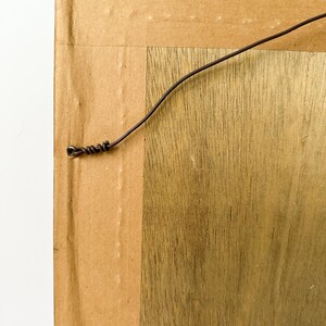 May include: Close-up of the back of a framed artwork. The frame is made of wood with a natural grain pattern. A dark wire is attached to the back for hanging. The backing is cardboard.