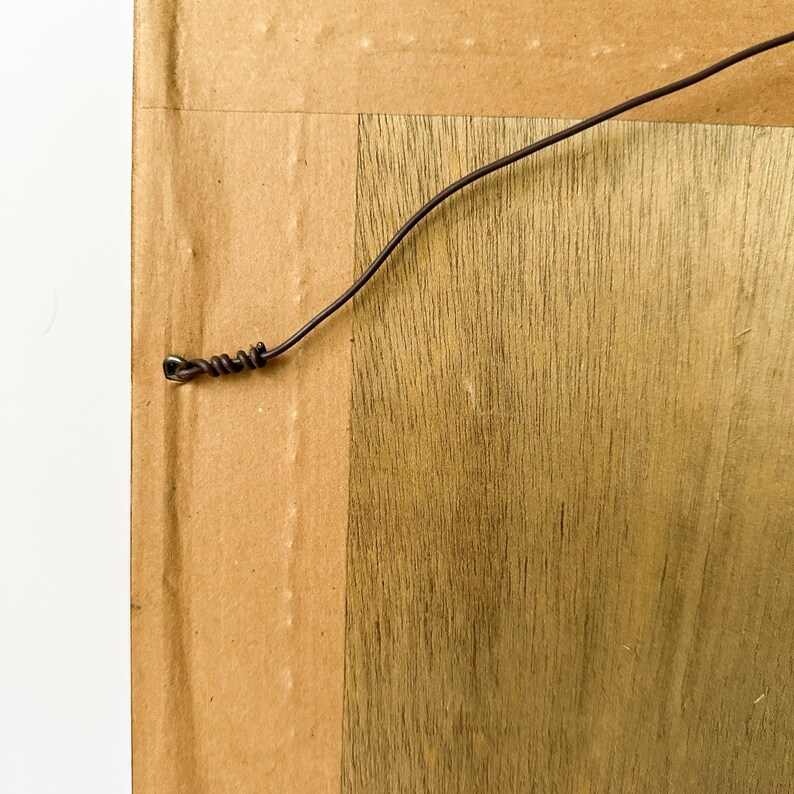 May include: Close-up of the back of a framed artwork. The frame is made of wood with a natural grain pattern. A dark wire is attached to the back for hanging. The backing is cardboard.