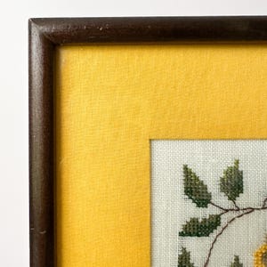 May include: A framed cross-stitch artwork featuring a floral design. The artwork is set against a yellow background and framed in a dark brown wooden frame. The cross-stitch depicts green leaves and yellow flowers.