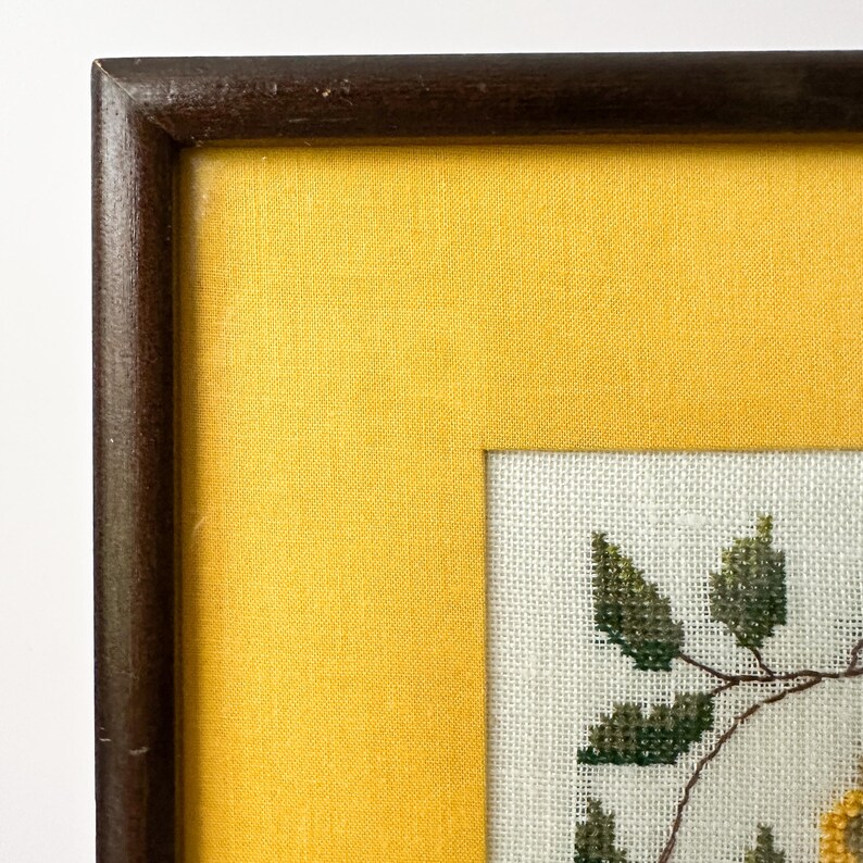 May include: A framed cross-stitch artwork featuring a floral design. The artwork is set against a yellow background and framed in a dark brown wooden frame. The cross-stitch depicts green leaves and yellow flowers.