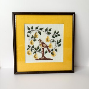 May include: Framed cross-stitch artwork featuring a bird perched on a tree branch laden with yellow pears. The embroidery is set against a cream-colored background, framed by a yellow mat and a dark brown wooden frame. A charming piece of home decor.
