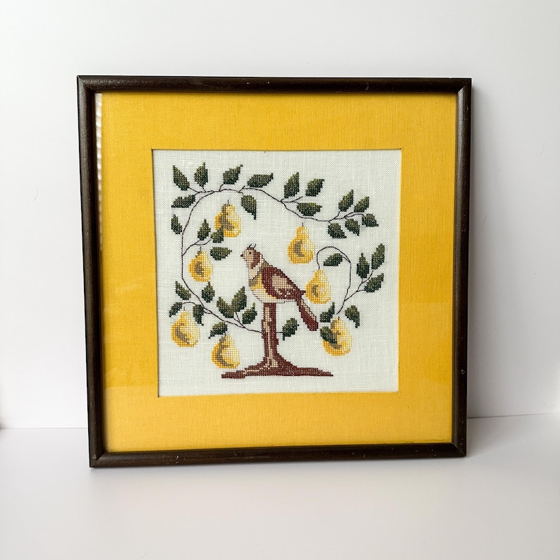 May include: Framed cross-stitch artwork featuring a bird perched on a tree branch laden with yellow pears. The embroidery is set against a cream-colored background, framed by a yellow mat and a dark brown wooden frame. A charming piece of home decor.