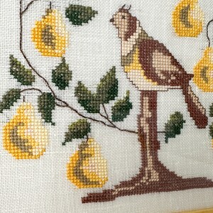 May include: Cross-stitch artwork featuring a bird perched on a tree branch with yellow pears. The bird has brown, tan, and yellow details. The pears and leaves are green and yellow. The background is white.