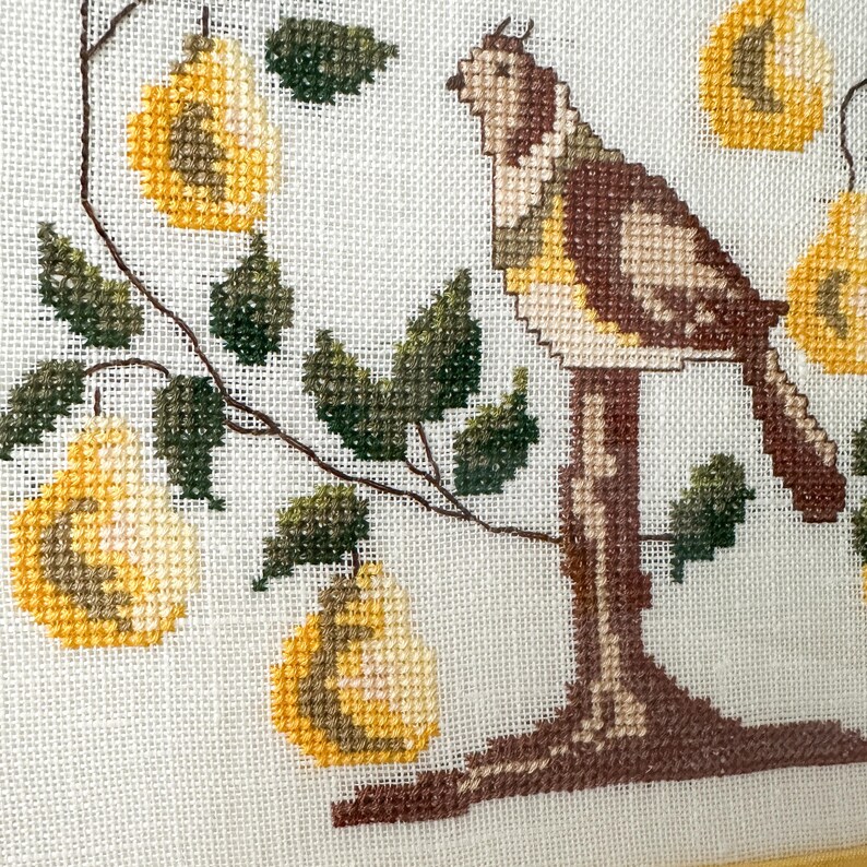 May include: Cross-stitch artwork featuring a bird perched on a tree branch with yellow pears. The bird has brown, tan, and yellow details. The pears and leaves are green and yellow. The background is white.