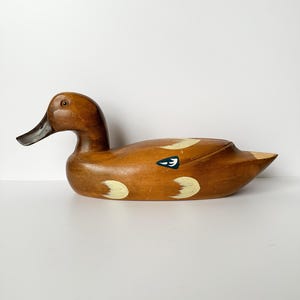 May include: A wooden duck decoy with a brown body, black beak, and painted white and yellow accents. The duck has a small blue and white painted detail on its side. The decoy is a decorative item.
